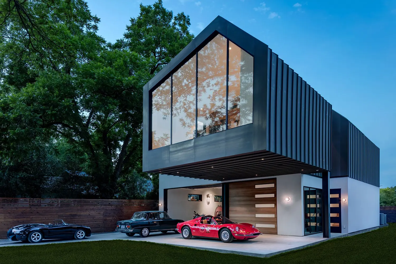 AutoHaus by Matt Fajkus Architecture, twilight exterior view of gabled steel-clad volume with three vintage cars parked below