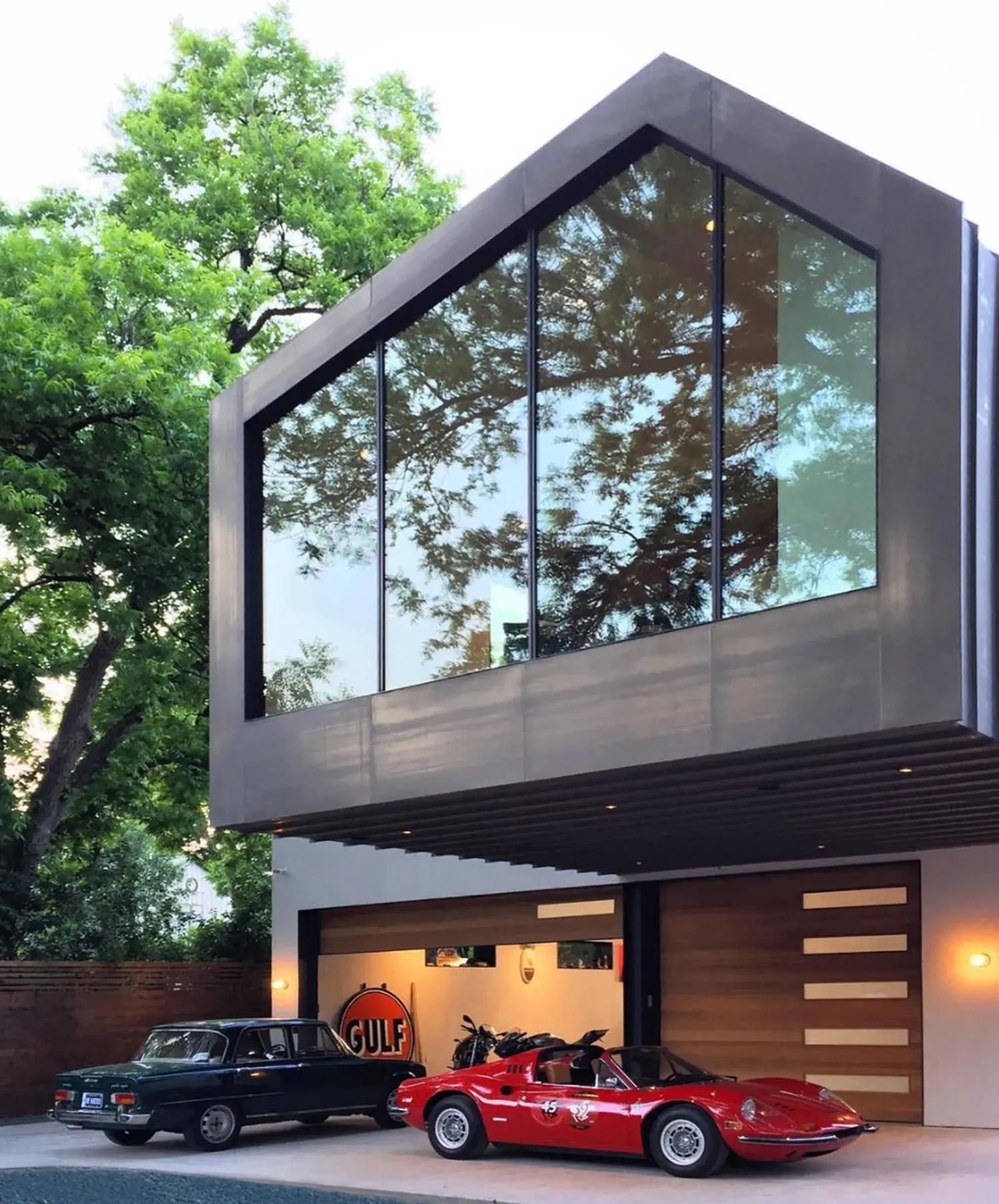 AutoHaus by Matt Fajkus Architecture, twilight exterior view of gabled glass facade with two vintage cars and Gulf sign visible