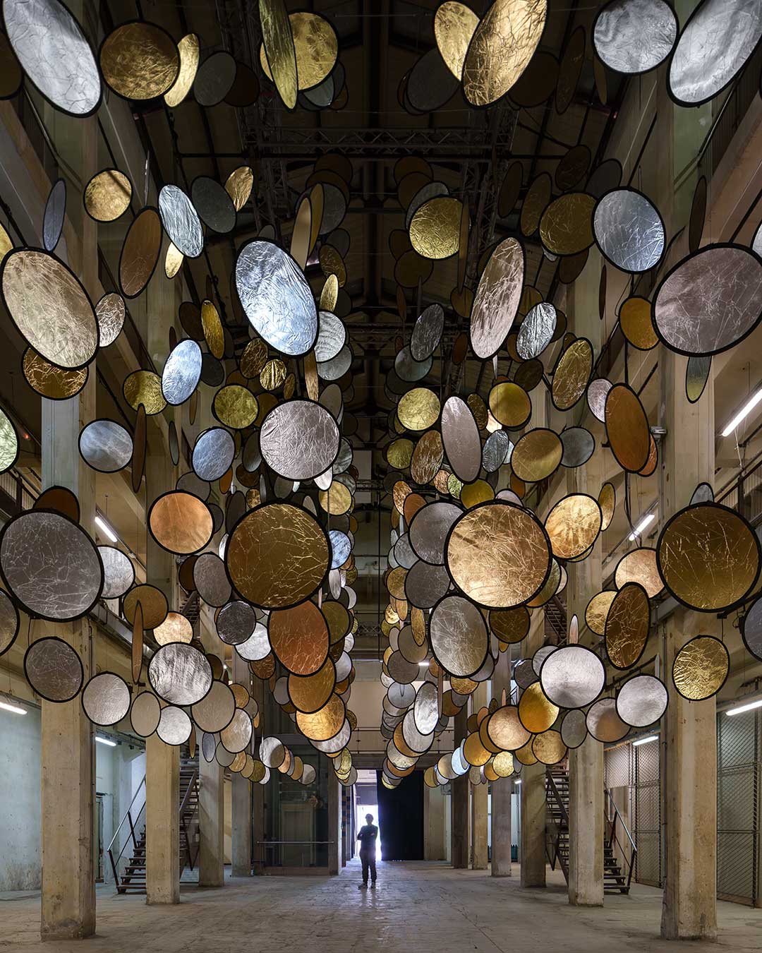 Suspended in the industrial void of a former Florence railway factory, HALOS acts as a living landscape of gold and silver reflections. Spanish artist SpY has introduced a rhythmic choreography of rotating discs that breathe through motion, catching ambient light to throw a fragmented spray of reflections across the building's skeletal frame. The atmosphere shifts between solar warmth and lunar coolness, creating a sensory experience defined by weightlessness and a profound state of suspension.  As visitors navigate the atrium’s three levels, the installation evolves from a suspended sky into a dense, topographical landscape of light. This mastery of engineering allows the technology to disappear, leaving only a "metallic heartbeat" that brings a temporary, shimmering life to the industrial memory of the site. It is a reminder that even the most rigid structures can be reimagined through the intelligent application of light and motion.  via #urdesignmag
artist @spy__studio 
commissioned by @brightfestival 
images @rubenpb  #SpY #KineticArt #LightInstallation #Florence #IndustrialDesign