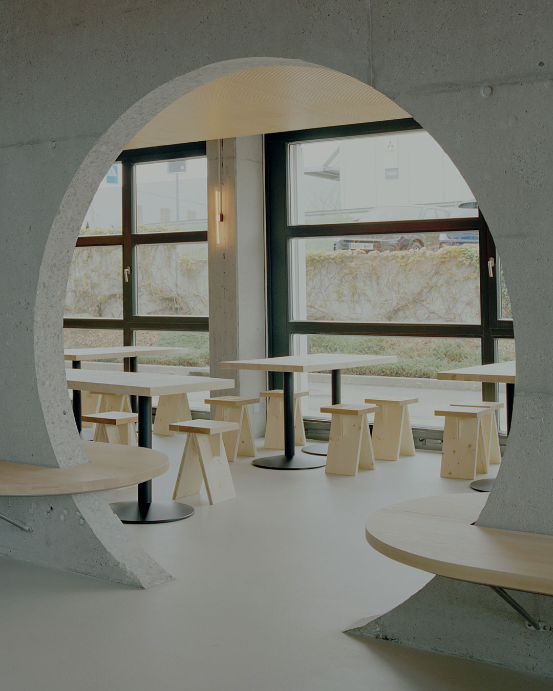 A load-bearing concrete wall in Granges-Paccot, Switzerland, couldn't be removed — so Noue Studio built the entire restaurant around it instead. One cutout through the wall does the work of a hallway, a sightline and a room divider at once, while brushed stainless steel and polycarbonate paneling frame the raw concrete without polishing it.  Reuse runs through every decision: former steps from the existing structure become stools, and the original tables are recovered and put back into use rather than replaced.  architects: @noue.studio 
photography: @willempab  urdesignmag.com — link in bio  #NoueStudio #SwissArchitecture #RestaurantDesign #AdaptiveReuse #ConcreteArchitecture