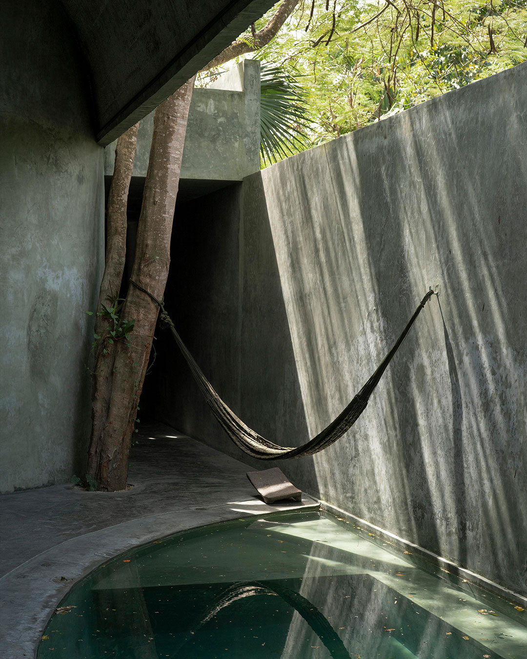A tree the architects refused to remove now grows straight through the entrance vestibule of @casa.gruta — the first sign that this Valladolid house was designed as a threshold, not a facade. Salvador Román and Adela Mortera cast the entire 254 sqm (2,734 sq ft) structure from pigmented concrete tuned to shift color with the sun, echoing the saturation of the region's limestone cenotes.  Inside, a vaulted, hammer-textured concrete ceiling leads from a compressed entry tunnel into a plunge pool and hammock corridor, while a private bedroom courtyard holds its own circular water feature. Brass pendant lighting and a reclaimed wood kitchen island are the only materials the architects allowed to depart from concrete.  architects: @chavaroman 
photography: @fabianml  urdesignmag.com — link in bio  #CasaGruta #Yucatan #ConcreteArchitecture #MexicanArchitecture #InteriorDesign