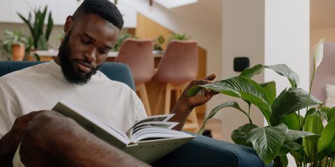 Man sitting on a sofa reading a book next to a plant
