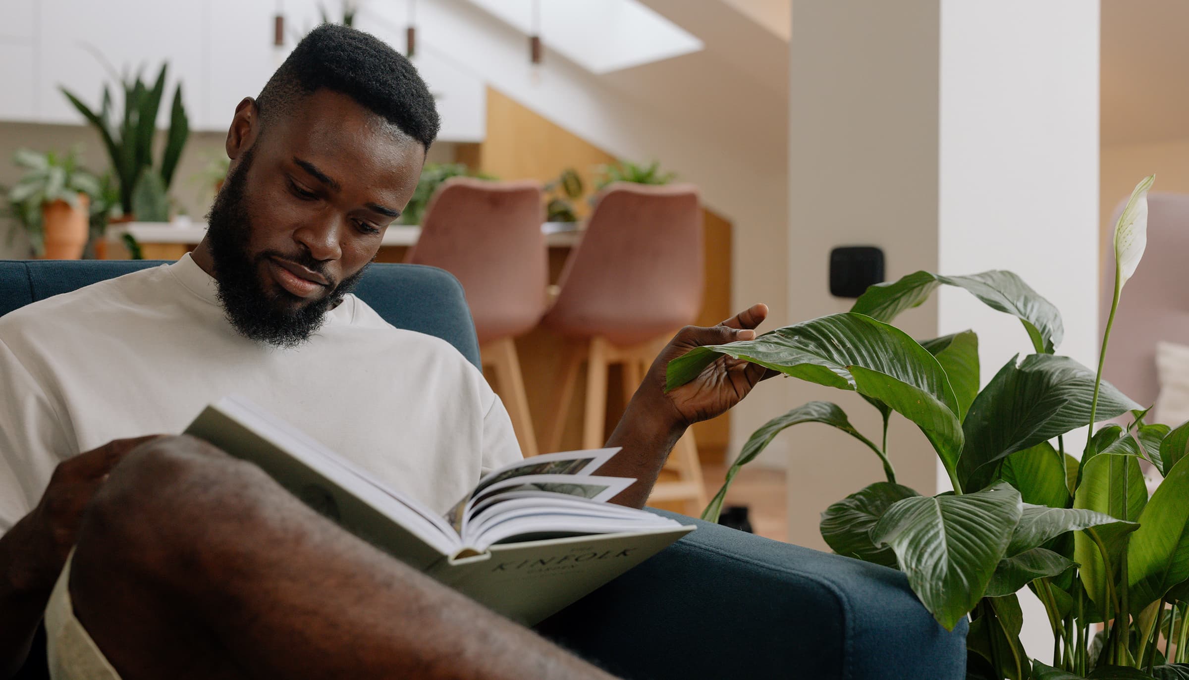 Man sitting on a sofa reading a book next to a plant