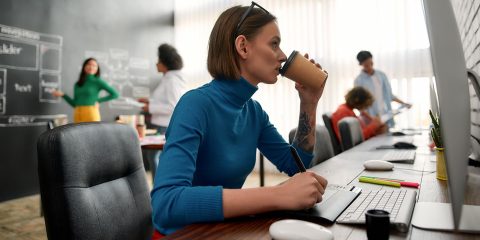 Side view of a young female web developer working on computer and drinking coffee in the creative office