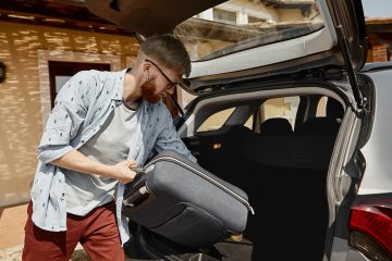Outdoor shot of handsome trendy looking young man with stubble holding suitcase while putting his luggage in trunk