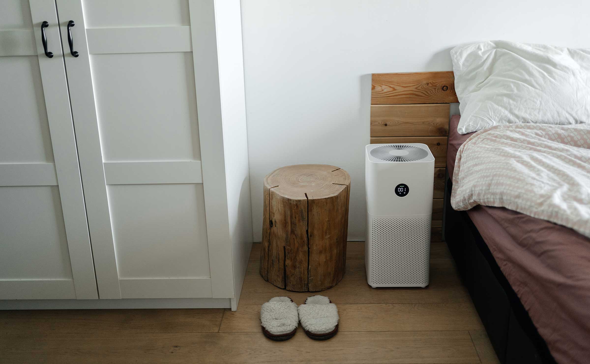 The air purifier stands on the floor in the bedroom next to the bed and cleans the air of dust