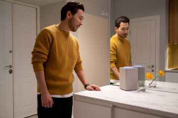 Photo of a Man in a Mustard Sweater Near a Mirror Interacting With Smart Speaker