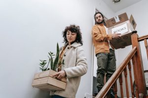 A Couple Moving Out Carrying A Box And Crate With Plants