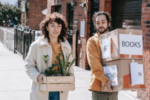 Couple Walking In The Street Carrying Plants And Boxes