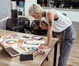 Young stylish blonde tattooed female designer making some sketches and working with color swatch samples while standing near office desk
