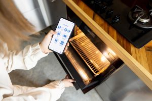 Woman holding phone with running smart home application, controlling kitchen devices remotely