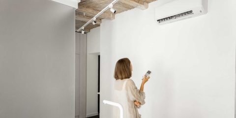 Woman sets the temperature on the air conditioner with remote control, while standing at modern white kitchen of the studio apartment