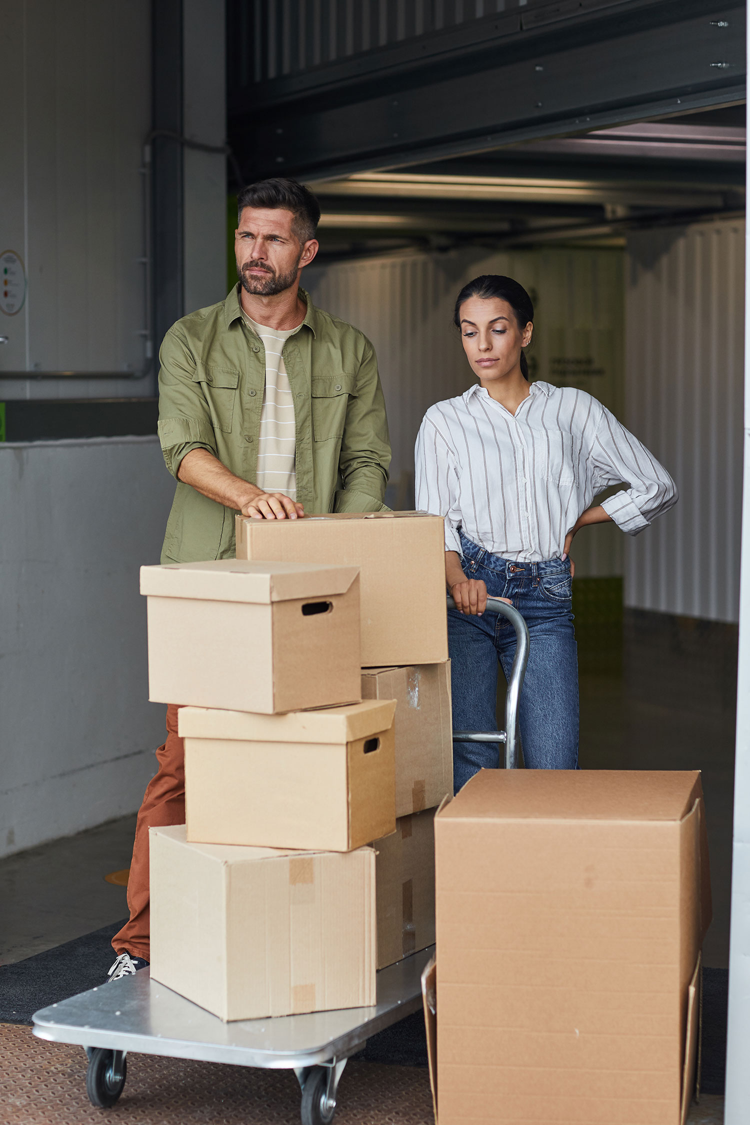 Vertical full length portrait of modern couple standing by cart with cardboard boxes while loading self storage unit