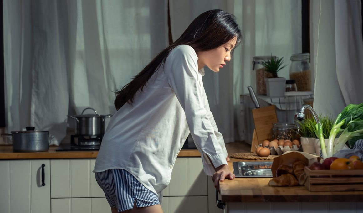 Depressed young beautiful Asian woman standing in the kitchen with stressed face.