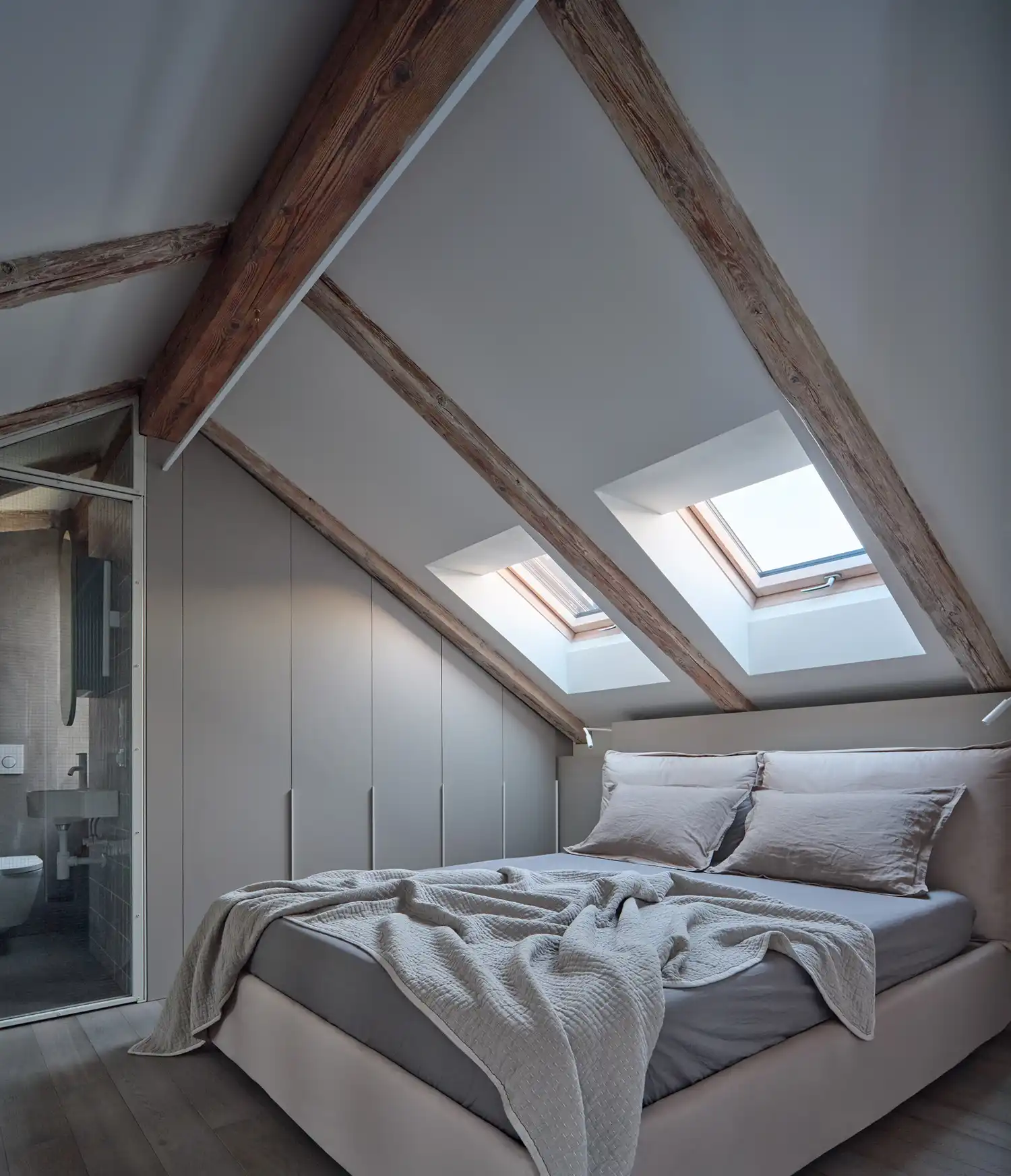 Airy loft bedroom in Formafatal’s Prague apartment, showcasing cream walls, exposed timber rafters, and a minimalist walnut-clad storage block.