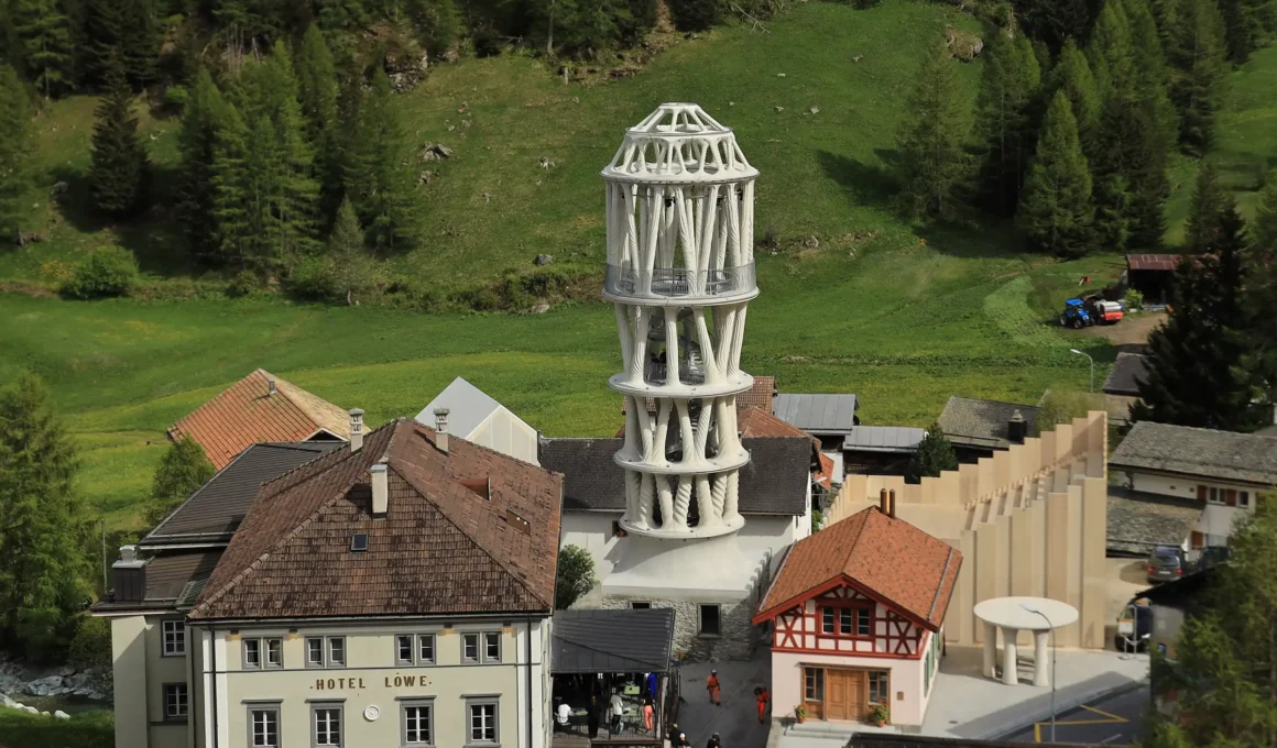 A towering 12-meter-high 3D-printed white concrete structure (Tor Alva) with intricate lattice-like patterns, standing against a Swiss alpine landscape in Mulegns.