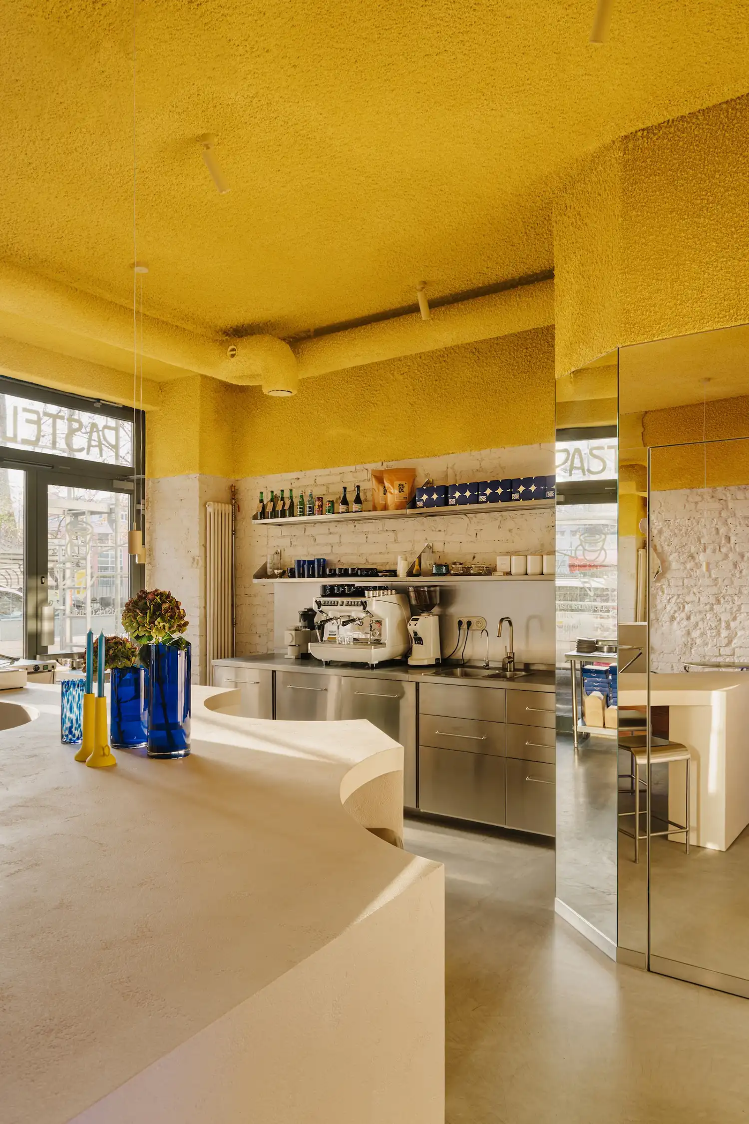 Undulating tunnel-like pastry counter at Pastelowe Warsaw, designed by Znamy Sie studio to mimic a reef-break wave. Glossy steel surface reflects coastal light.