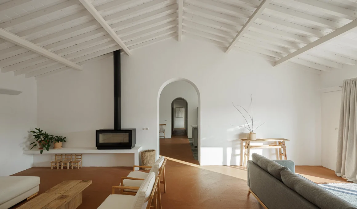Exterior view of MONTE DO DIVOR in Estremoz, Portugal: preserved rectangular house with gabled roof, two chimneys, modern minimalist sliding glass doors, and a compacted earth path leading to the pool area, designed by Vasco Burnay Arquitectura.