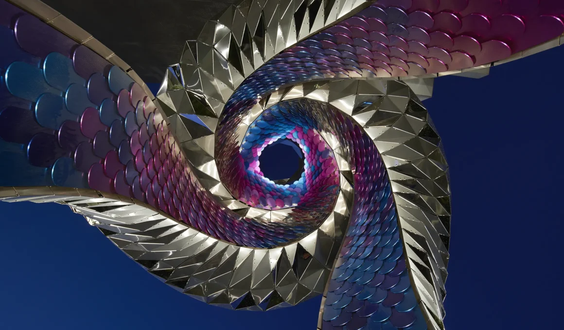 Close-up of FUTUREFORMS' Orbital sculpture at night, showing the swirling purple/blue interior shingles and engineered metal exterior.