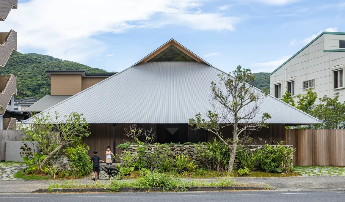 An off-grid Japanese residence by Sakai Architects on Amami Island featuring a large, silver corrugated-metal irimoya roof and integrated lush gardens behind a traditional stone wall.