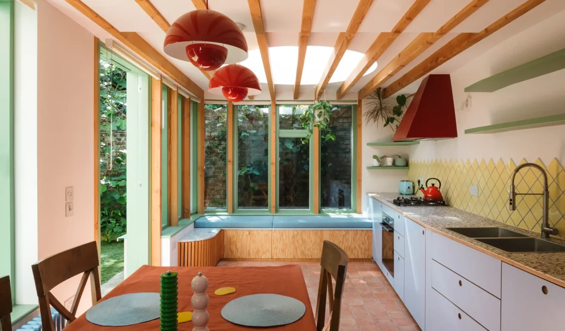 Interior view of a vibrant, light-filled kitchen extension by Office S&M featuring exposed timber beams, a circular skylight, light blue cabinetry, and bold red and yellow accents.
