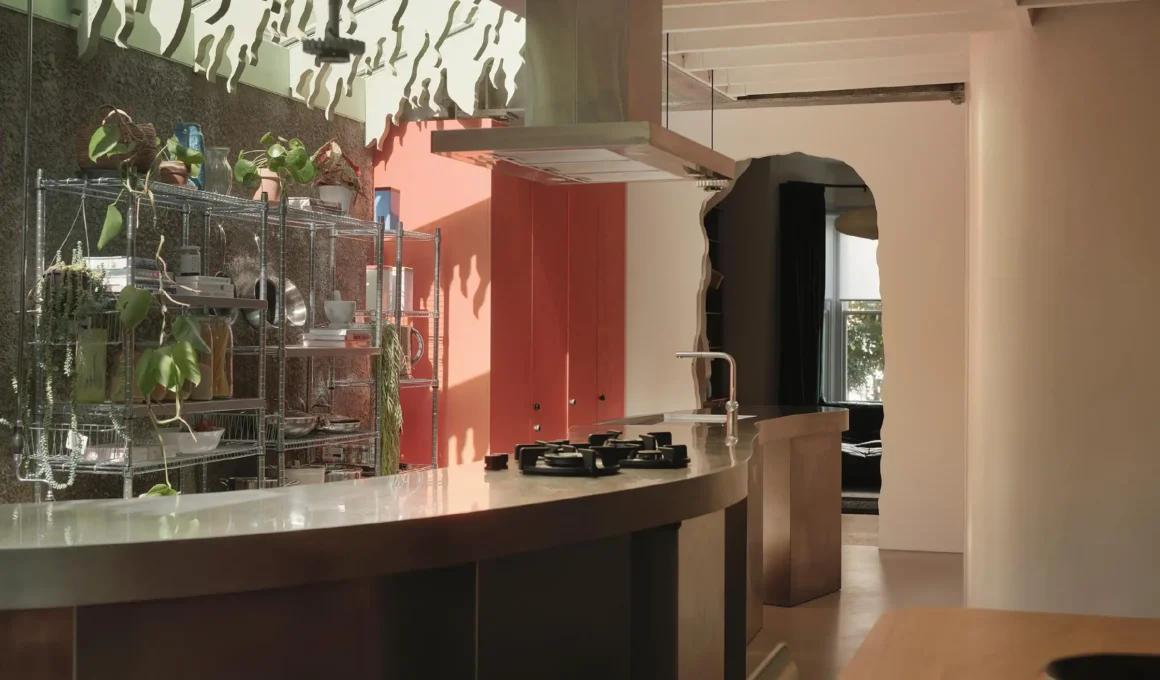 Organic, cave-like archway in a white room leading into the textured, material-rich kitchen area with a stainless steel island and green ceiling feature in CAN's Druid Grove House, London.