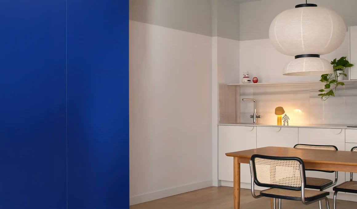 A modern open-plan dining area in Valencia featuring a bold Klein blue storage volume, white Macael marble kitchen backsplash, and historic white ceiling moldings above a contemporary wooden dining set.