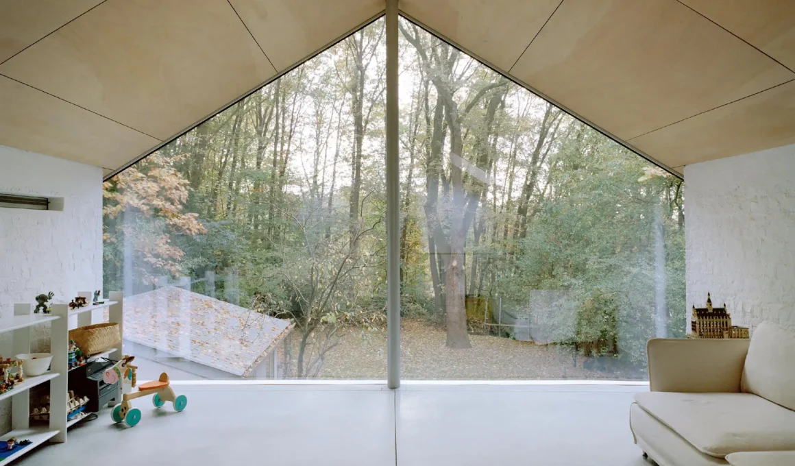 Elmēs architecture attic living space in Le Regain House Overijse, Belgium featuring minimalist plywood ceiling, polished concrete floor, and full-height triangular glass façade overlooking a protected forest, showcasing modern Belgian design and natural light.