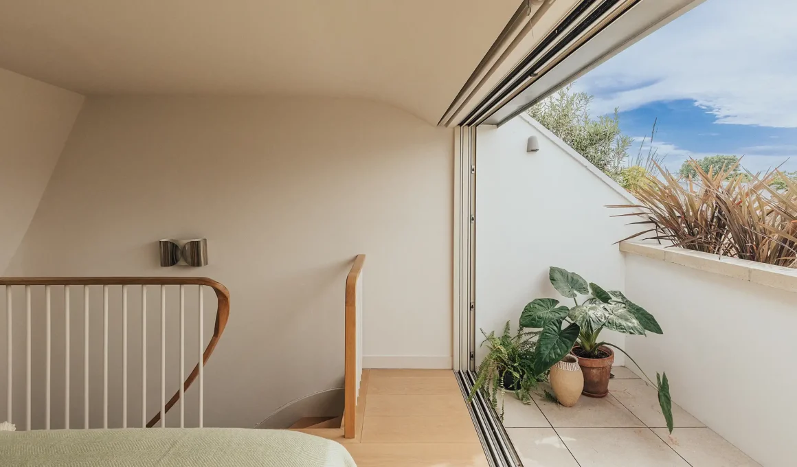 A minimalist contemporary bedroom by Emil Eve Architects featuring floor-to-ceiling bespoke oak wardrobes, a mid-century wooden armchair, and natural light streaming onto wide oak floorboards.