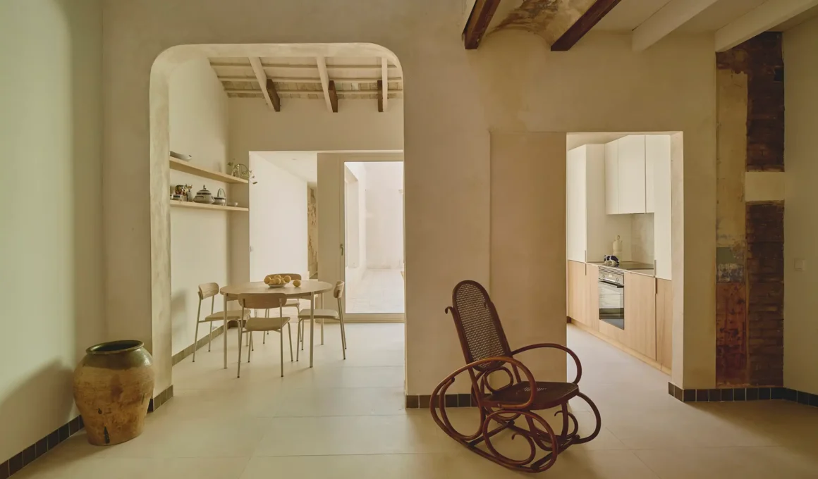 Interior of Estels House by ENDALT Arquitectes featuring a minimalist living area with a traditional rocking chair, original timber beams, and a view into the kitchen and dining space.