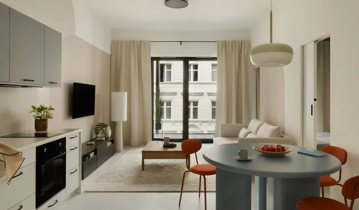 A modern, minimalist European apartment interior featuring a compact open-plan kitchen and living area, looking out onto a traditional urban streetscape through a large window.