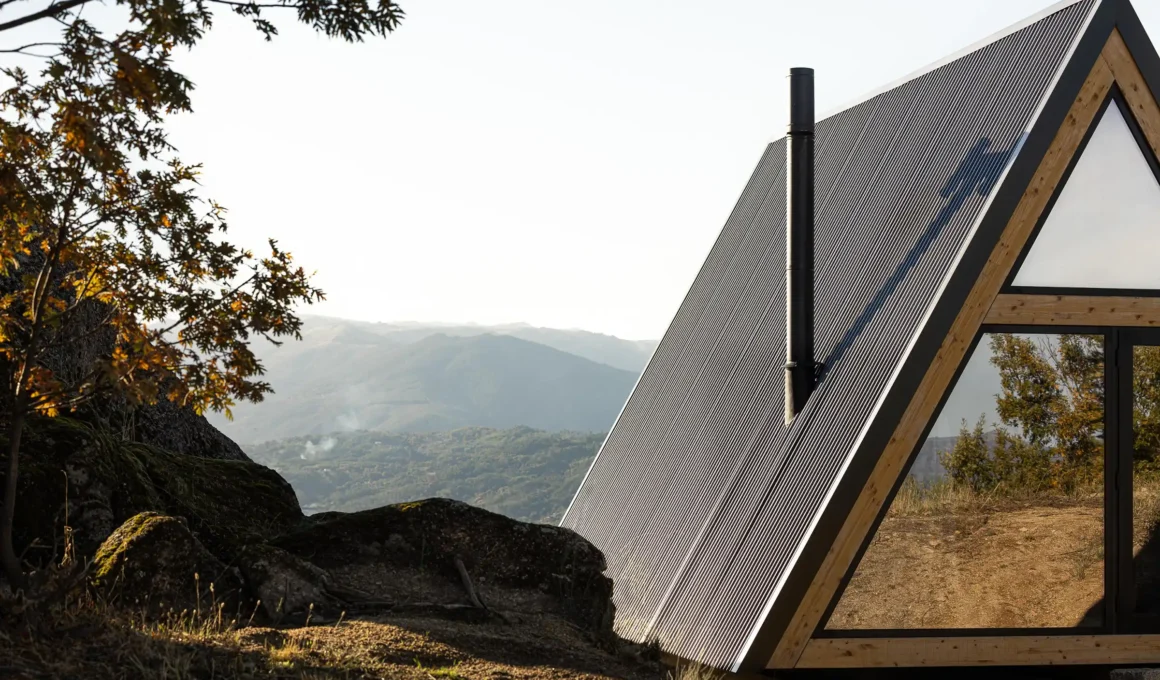 Minimalist triangular mountain shelter by Filipe Pina Arquitectura, finished in black corrugated metal and wood, set on rocky terrain with a fully glazed facade overlooking a valley in Serra da Esperança, Portugal.