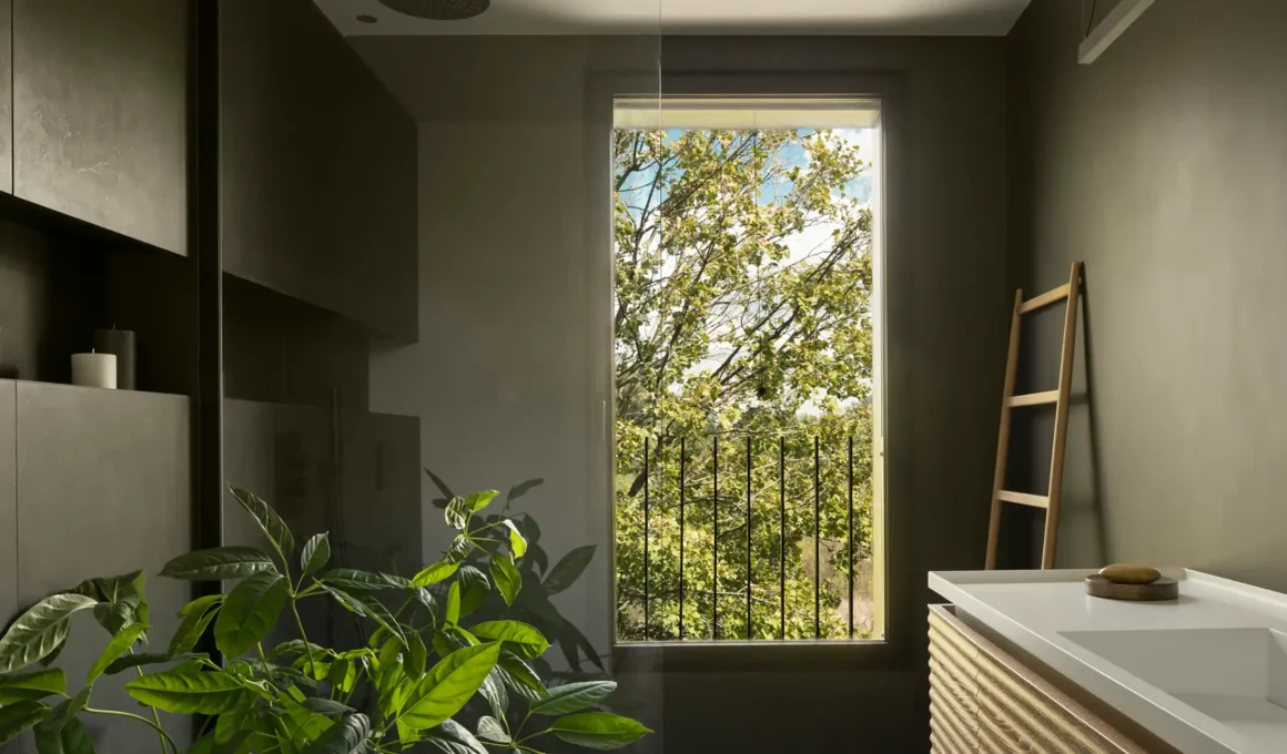 Minimalist bathroom in the Pivot House by Locus Architetti featuring deep green walls, a fluted timber vanity, and a large window framing a tree.