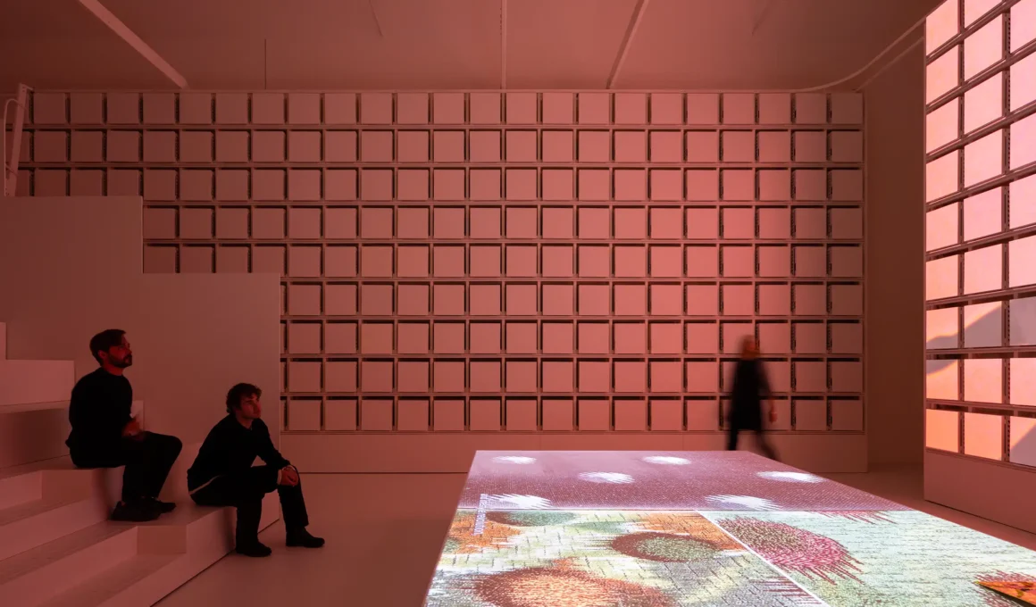 An interior view of the MAE Museum designed by CRA-Carlo Ratti Associati and Italo Rota, featuring a gridded wall of archival boxes and an interactive digital floor projection in a warm-toned gallery.
