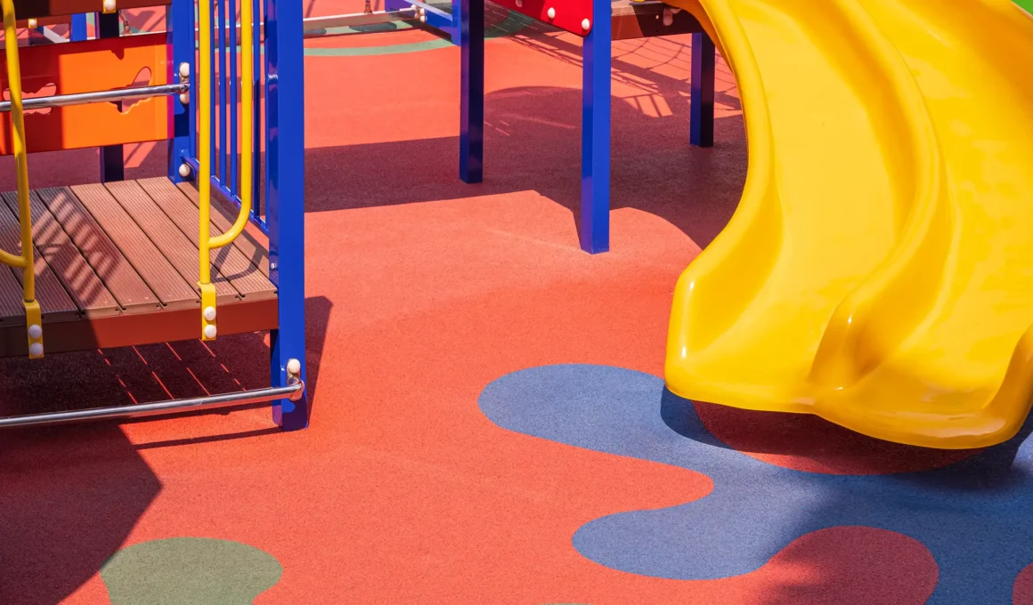 Modern playground safety surfacing with yellow slide and colorful, inclusive play equipment for children.
