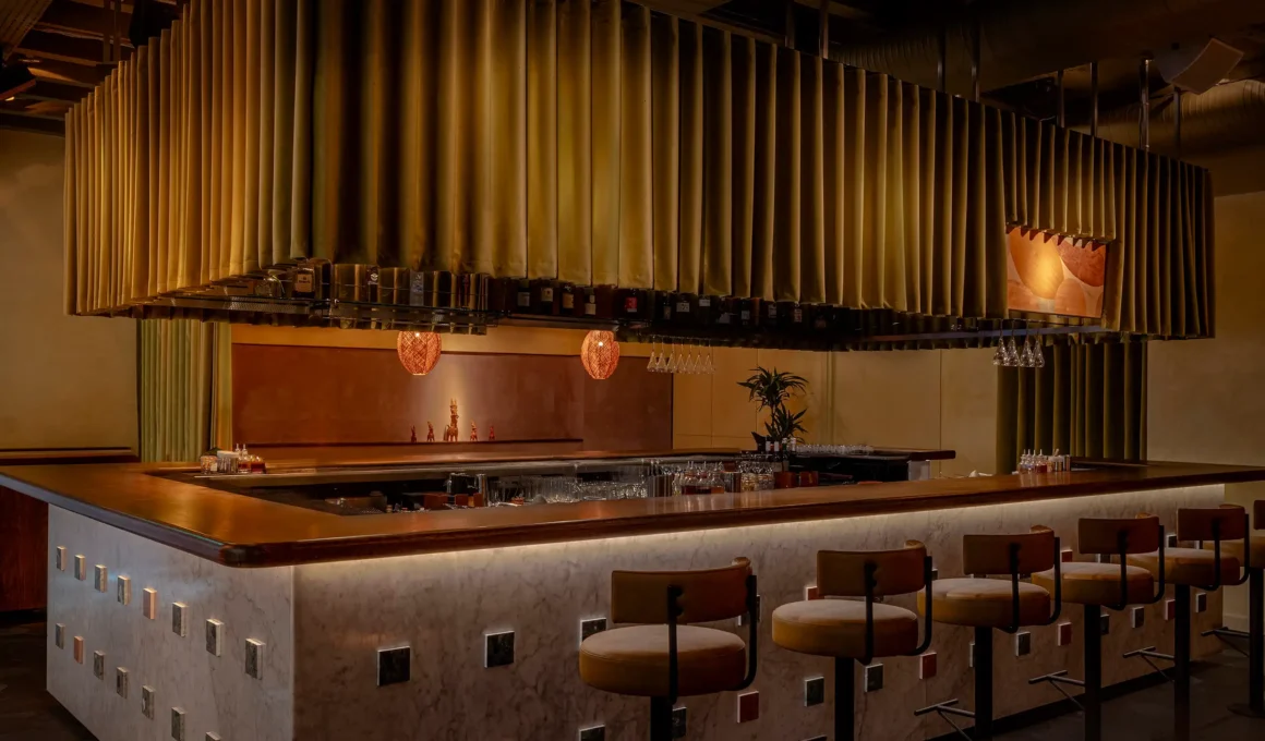 Monolithic marble cocktail bar with dark oak countertop and green velvet suspended curtain in London.