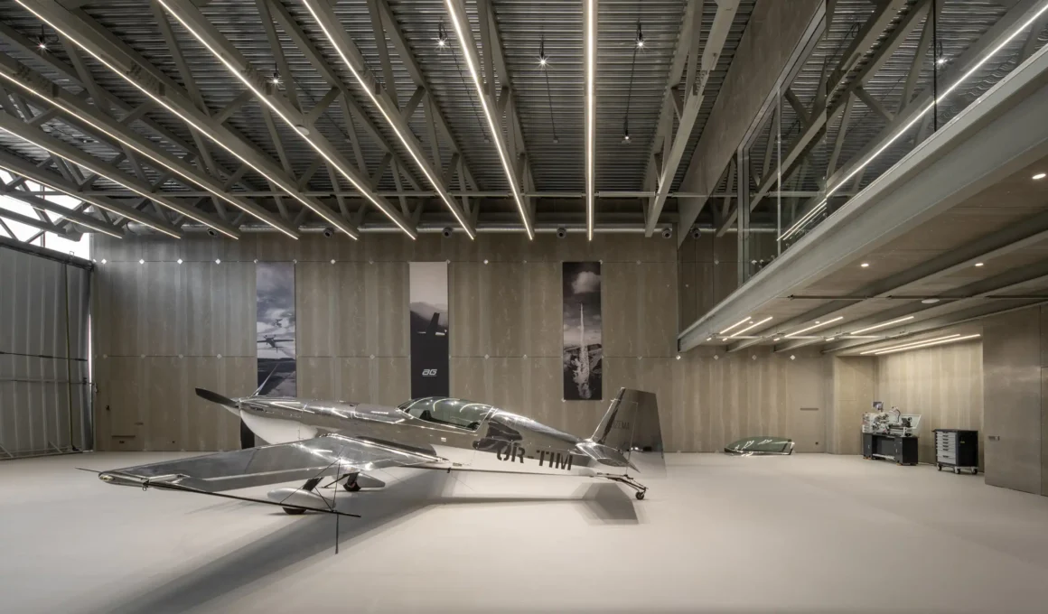 Wide-angle interior of the AEROTIM hangar hall featuring a silver aircraft, exposed steel trusses, and linear LED lighting.