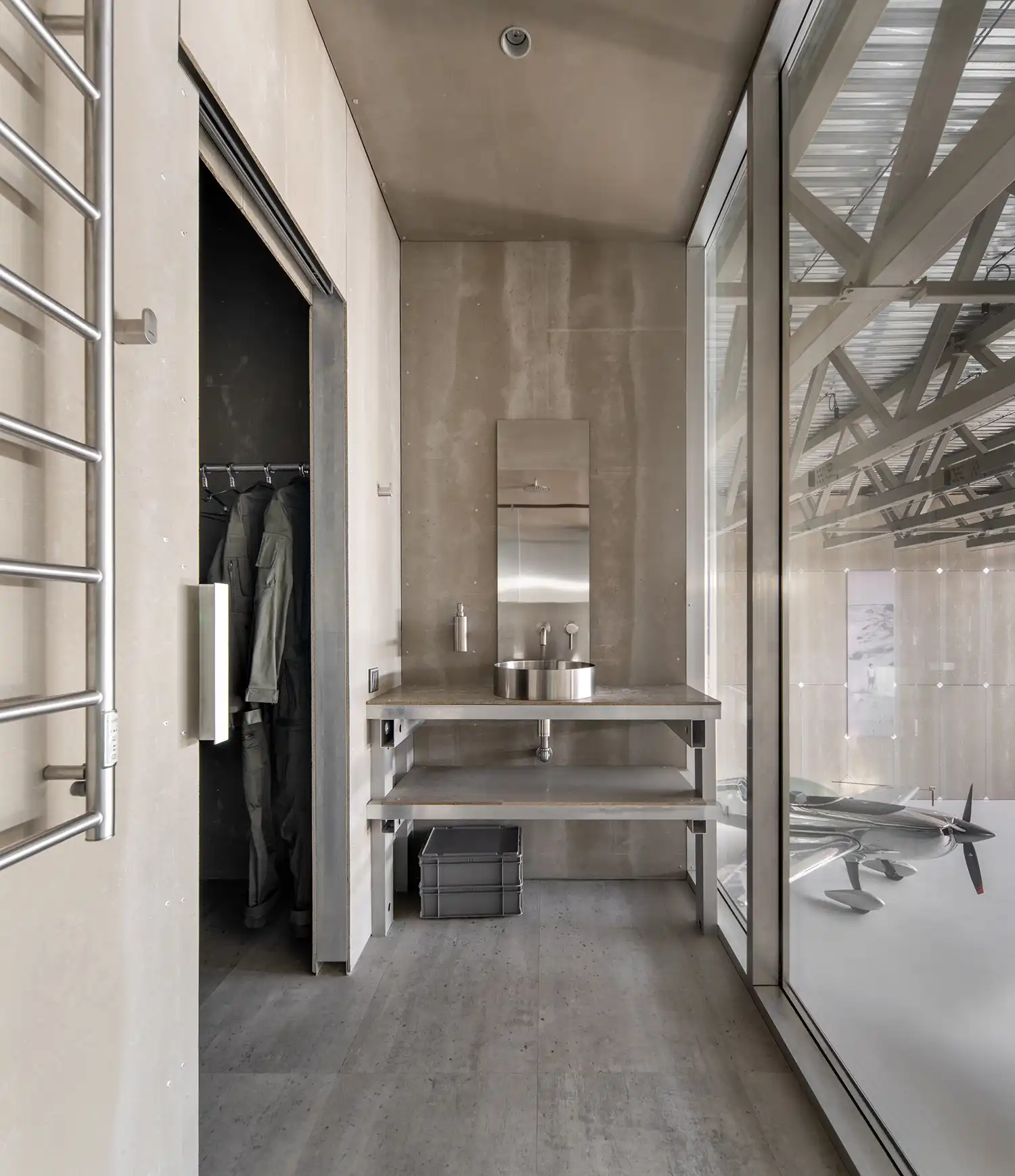 Minimalist changing room and shower area featuring a stainless steel sink, aluminum benches, and a glass window looking into the hangar.