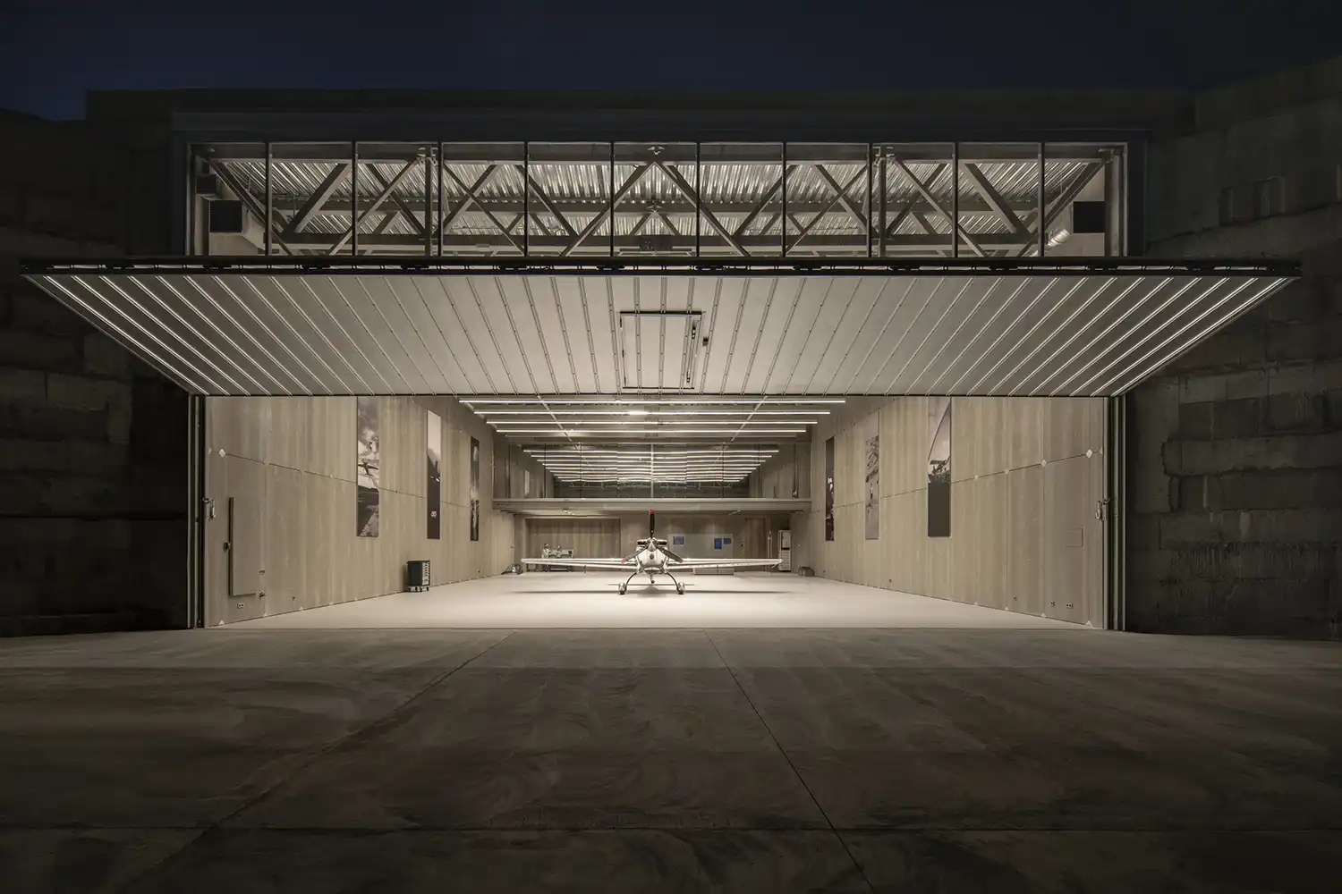 Exterior night view of the hangar with its large vertical gate fully open, revealing a brightly lit interior and a central aircraft.