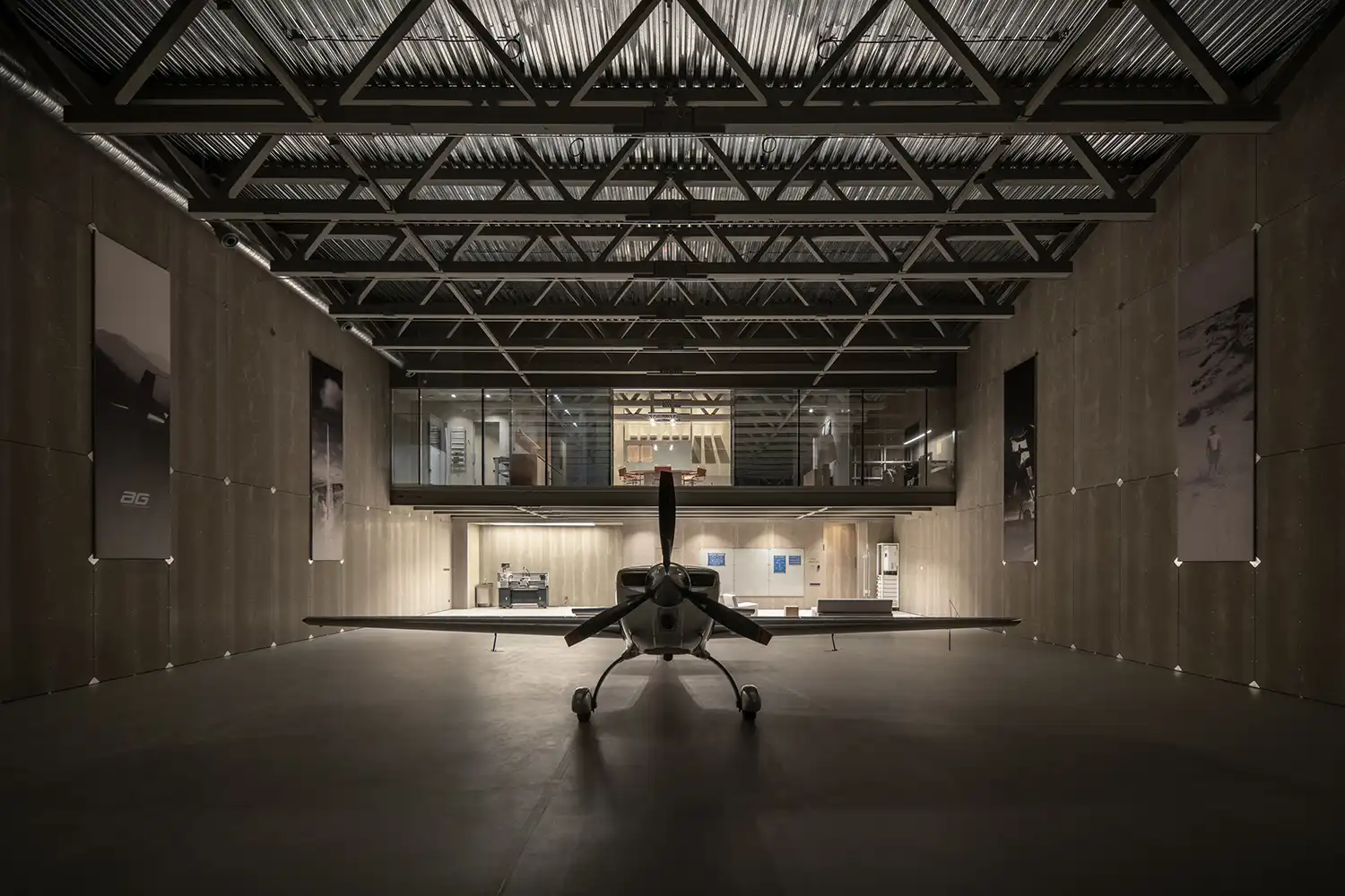 Symmetry-focused view from the back of the hangar hall looking toward the glass-fronted Crew Station with a silver aircraft in the foreground.