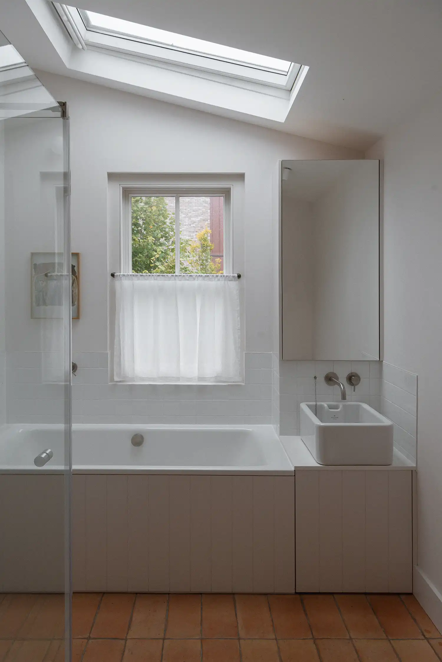 A minimalist bathroom with a vaulted ceiling, skylight, and red clay tile flooring by Architecture for London.