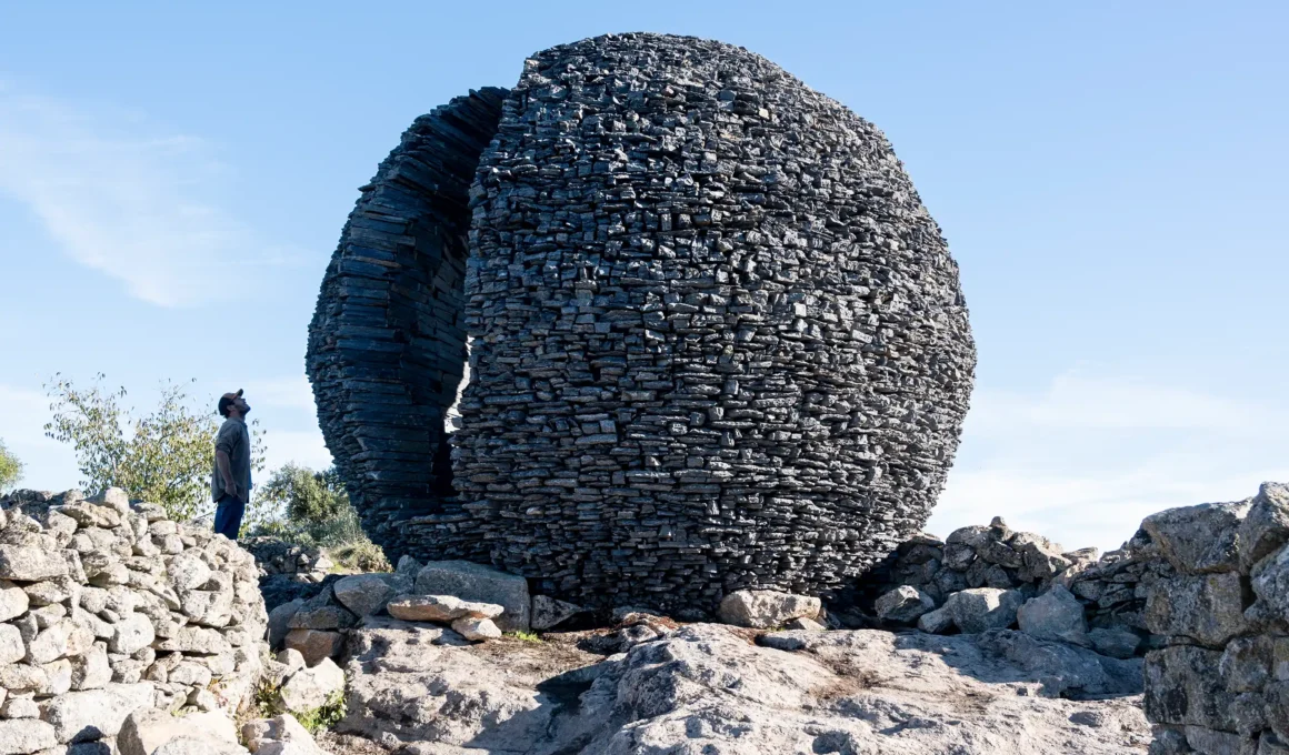 A large, spherical sculpture made of stacked black stone layers sitting on a rocky plateau under a clear sky.