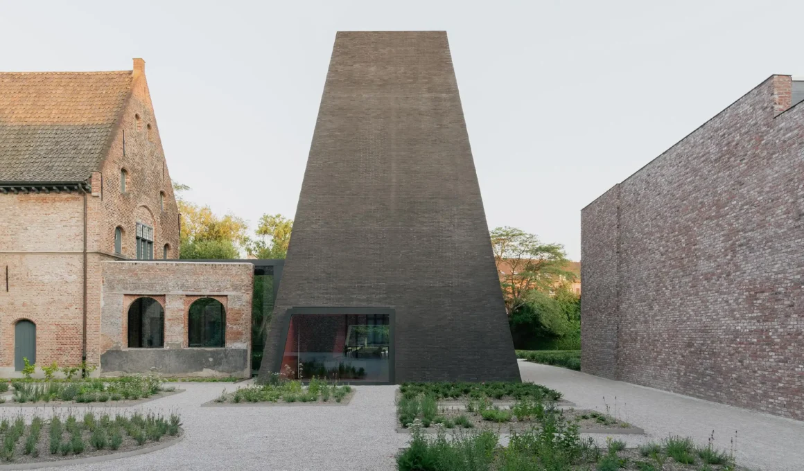A low-angle exterior shot of the Abby Kortrijk museum in Belgium featuring a dark, monolithic brick pavilion designed by Barozzi Veiga adjacent to a historic 16th-century brick abbey.