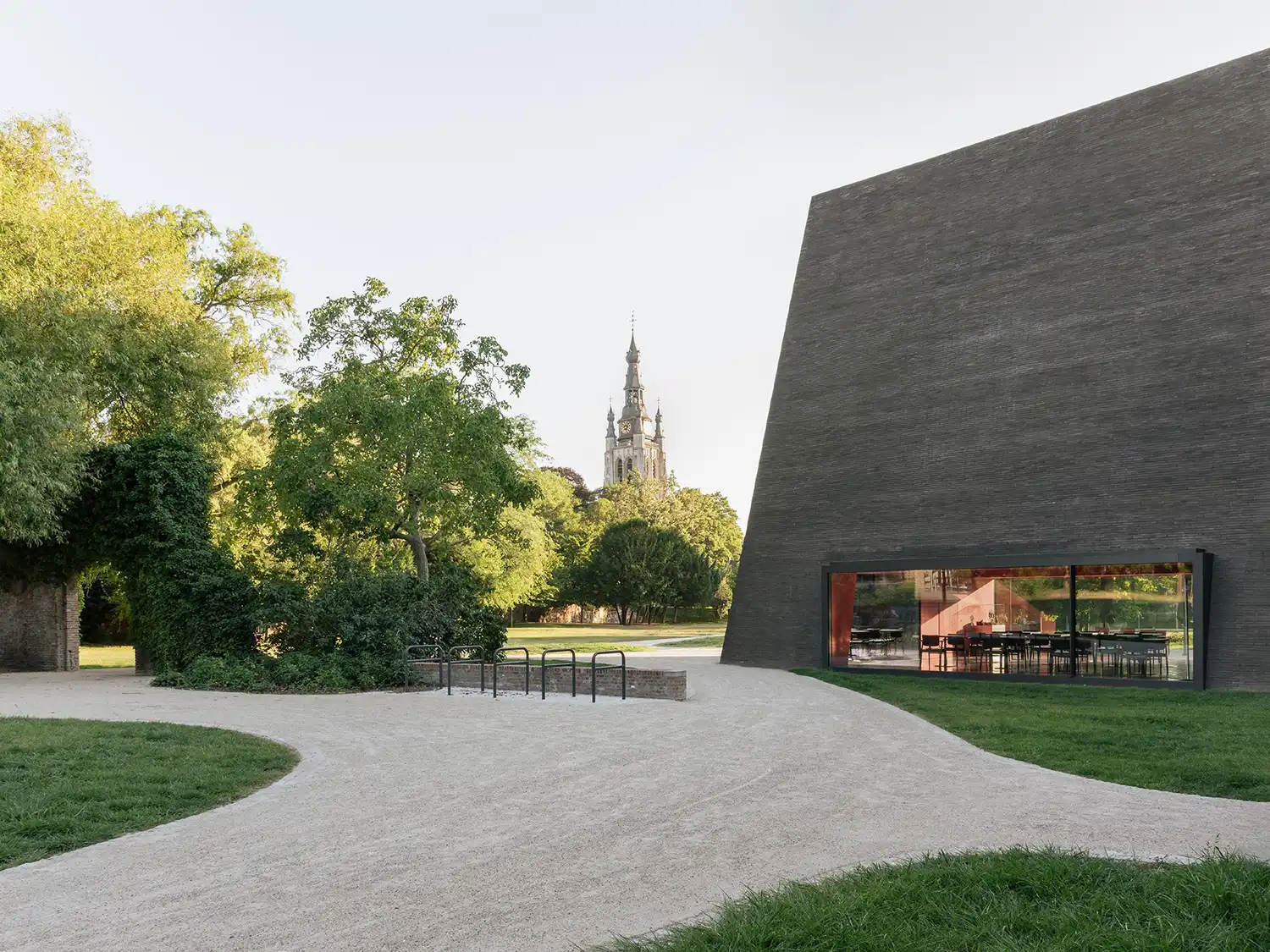 View of the Abby Kortrijk dark brick pavilion from a gravel public path, showing a large glass window and a church spire in the distant background under a clear sky.