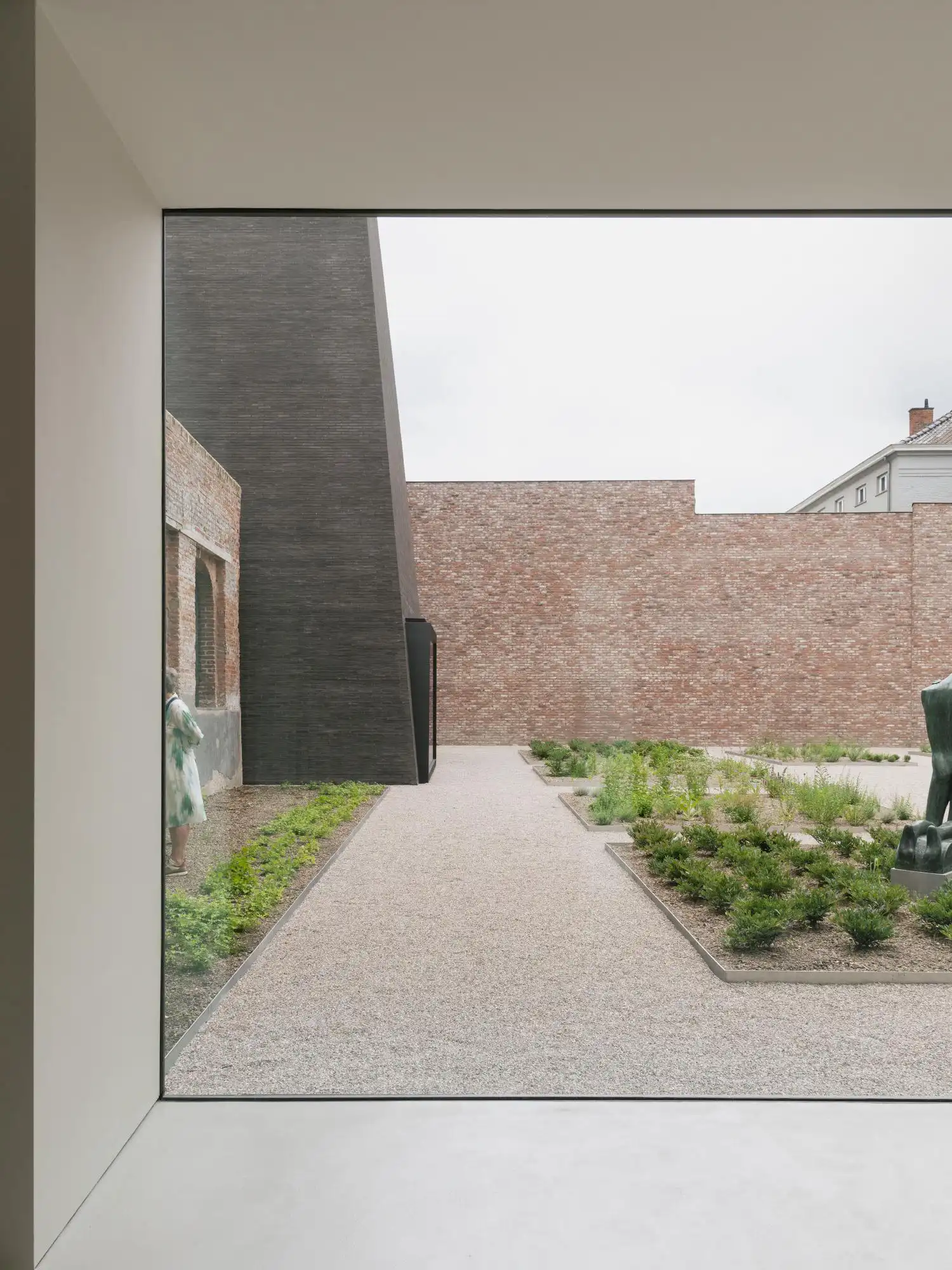 View from inside a white minimalist museum interior looking through a large floor-to-ceiling window at a landscaped courtyard and the dark pavilion of Abby Kortrijk.