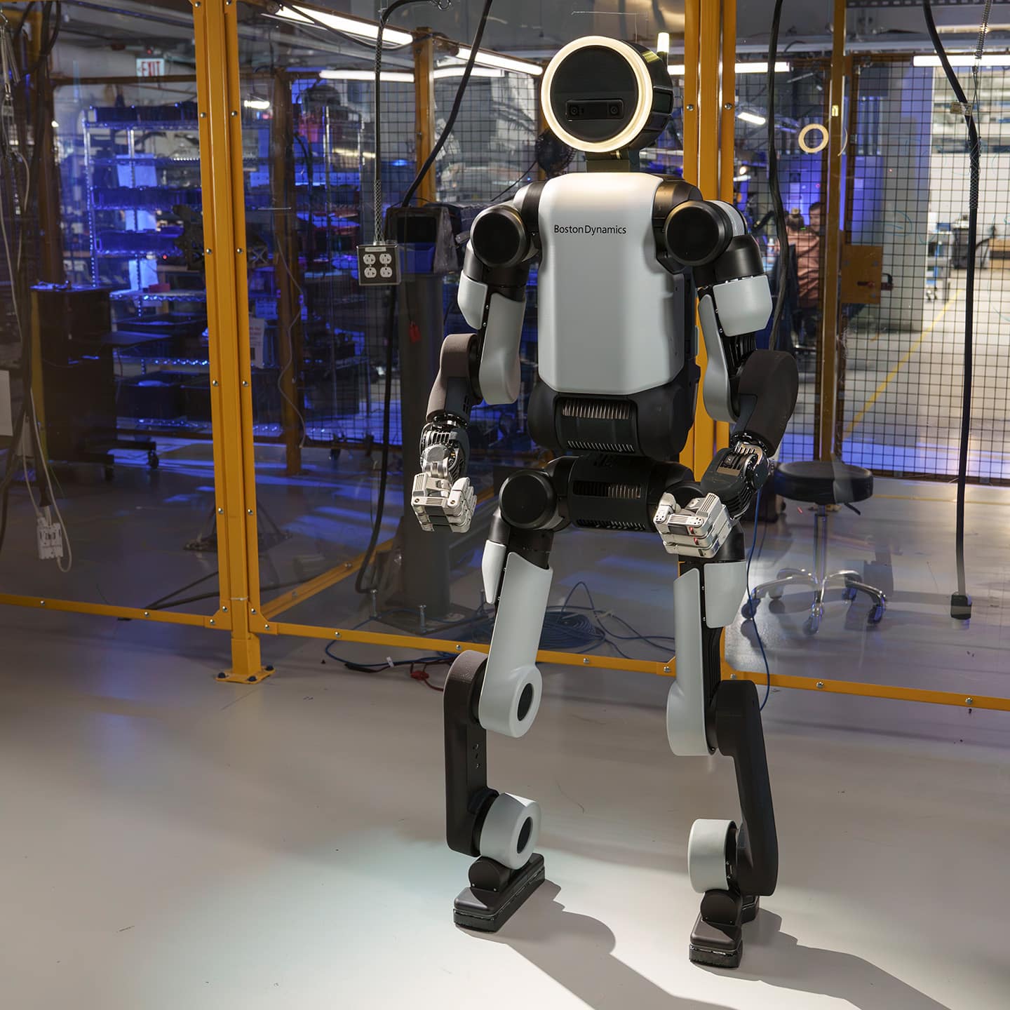 A full-body view of the Atlas robot featuring a white and black chassis standing within a glass-enclosed testing facility.