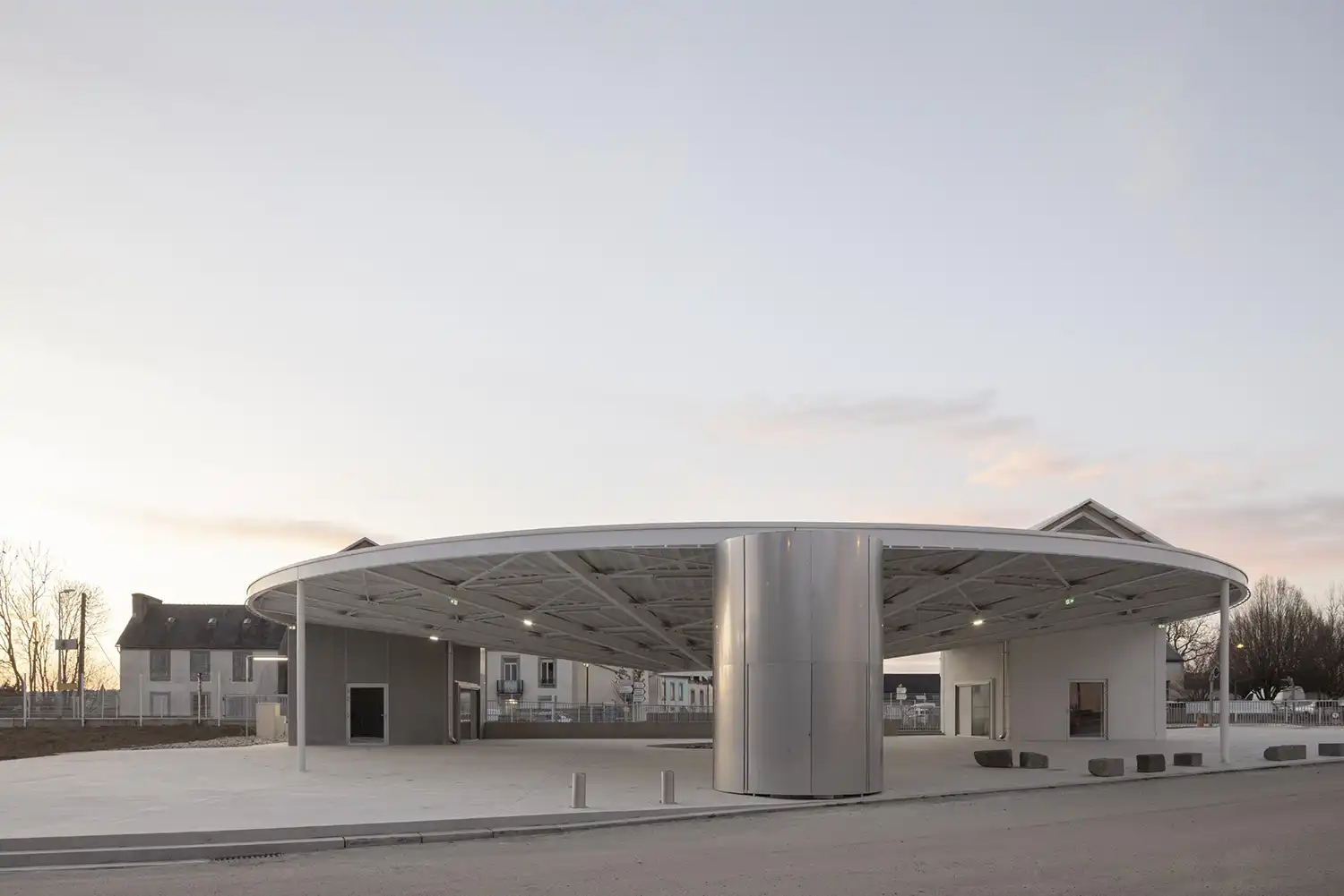 Exterior dusk view of the circular pavilion with integrated lighting and a prominent metallic technical shaft.