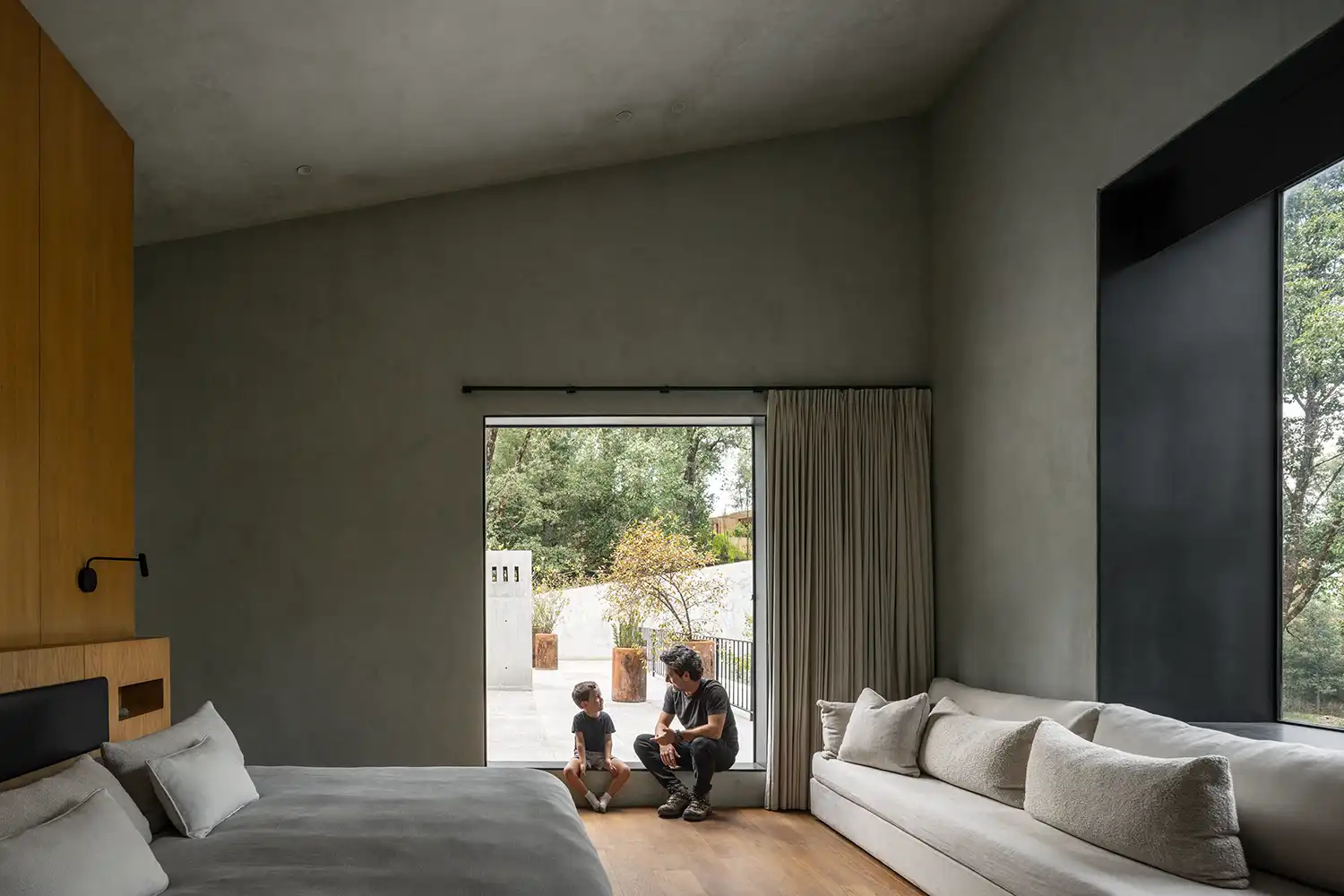 Minimalist bedroom with grey plaster walls and a large window looking onto a concrete patio.