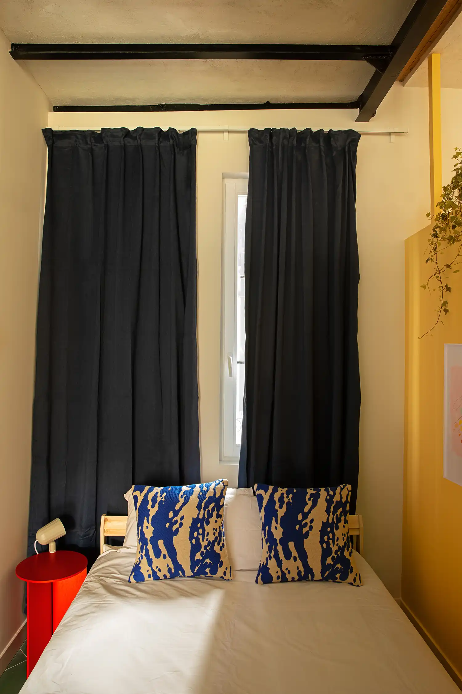 Small bedroom with white bed, blue patterned pillows, and a narrow window flanked by dark blue curtains.
