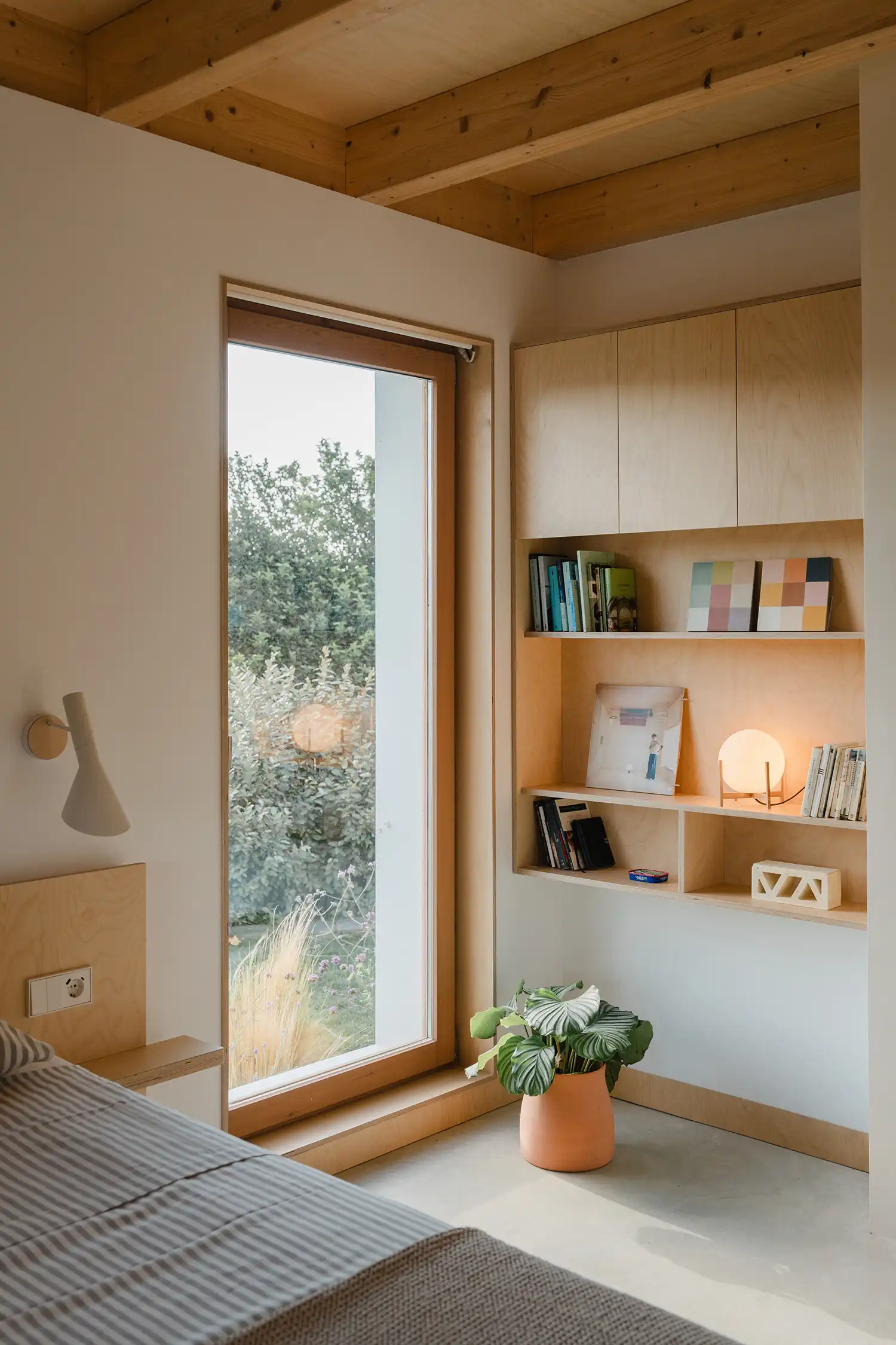 Cozy bedroom corner with floor-to-ceiling window and integrated plywood shelving units.